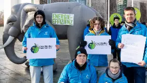 Fünf Menschen in blauen Jacken präsentieren im Freien das Logo des Umweltinstituts; hinter ihnen die Skulptur eines Elefanten, darauf steht «Energieeffizienz».