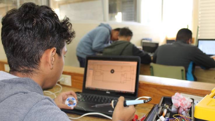 Ein junger Mann mit Lötzinn und -kolben in den Händen sitzt vor einem Laptop, auf dem ein Platinenschaltplan angezeigt wird. 