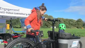 Vor einem offenen Infopavillon im Freien eine Frau in roter Jacke neben einem Lastenrad, in dessen Transportbehälter ein Kind mit Sicherheitshelm sitzt