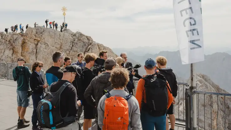 Menschen in Wanderkleidung aus einer Plattform unterhalb eines Gipfels, auch auf dem Gipfelgrat darüber reihen sich Menschen.