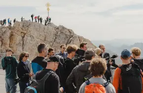 Menschen in Wanderkleidung aus einer Plattform unterhalb eines Gipfels, auch auf dem Gipfelgrat darüber reihen sich Menschen.