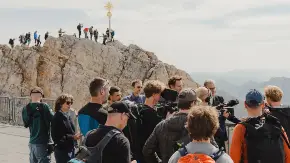 Menschen in Wanderkleidung aus einer Plattform unterhalb eines Gipfels, auch auf dem Gipfelgrat darüber reihen sich Menschen.