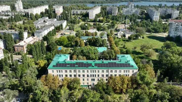 Luftaufnahme: Ein Krankenhaus, umgeben von einer Grünfläche und umrundet von mehrgeschossigen Wohngebäuden. Auf dem Dach sind PV-Module installiert.