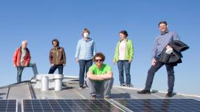 Eine Gruppe von zehn Menschen steht bei strahlendem Sonnenschein auf dem einem leicht geneigten Blechdach, im Vordergrund zahlreiche PV-Module.