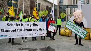 Vor dem Kanzleramt in Berlin präsentieren Aktivistinnen ein Banner, das Olaf Scholz auffordert, Atomkraft und Gas keinen grünen Stempael zu verleihen; rechts im Bild ein einzeln stehender Mensch mit übergroßer Olaf-Scholz-Maske, der ein «Nachhaltig»-Schild in der Hand hält.