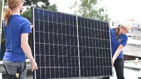 Zwei junge Menschen auf einem Hausdach tragen behutsam ein großes Photovoltaikmodul in Richtung eines im Hintergrund knienden Menschen.