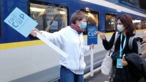 An einem regennassen Bahnsteig vor einem bereitstehenden Schnellzug weist eine Frau mit einem Schild «Rail to the COP» einer anderen Frau den Weg.