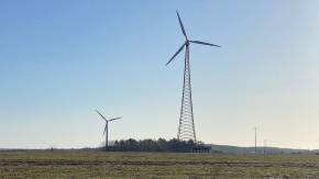 Zwei Windkraftwerke auf einer großen Wiese, im Hintergrund ein Wäldchen