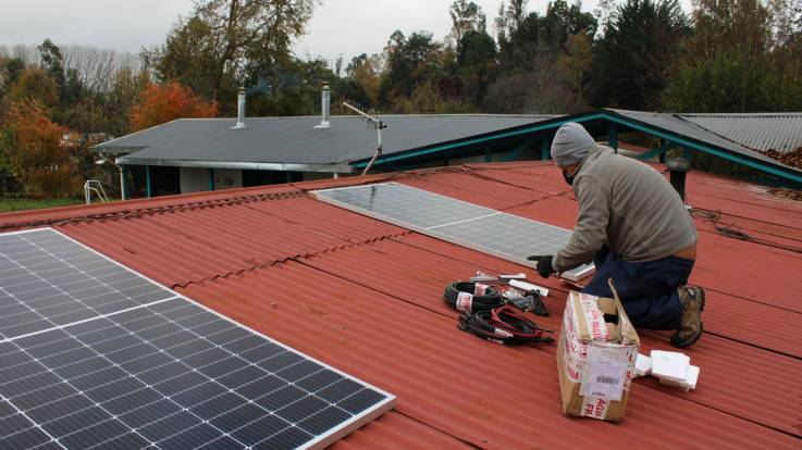 Auf einem roten Wellblechdach sind zwei PV-Module zur Montage ausgelegt, vor ihnen kniet ein Mann, neben ihm liegen Kabel, Befestigungsschienen sowie ein geöffneter Versandkarton mit weiteren Zubehörteilen.