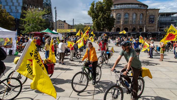 Teilnehmende an einer Fahrraddemo auf dem Platz vor dem Theater Freiburg, sie haben viele Flaggen mit der Anti-Atom-Sonne dabei.